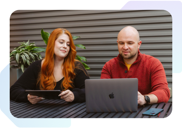 A woman with a tablet and a man with a laptop sitting next to each other at a table looking at the screen.