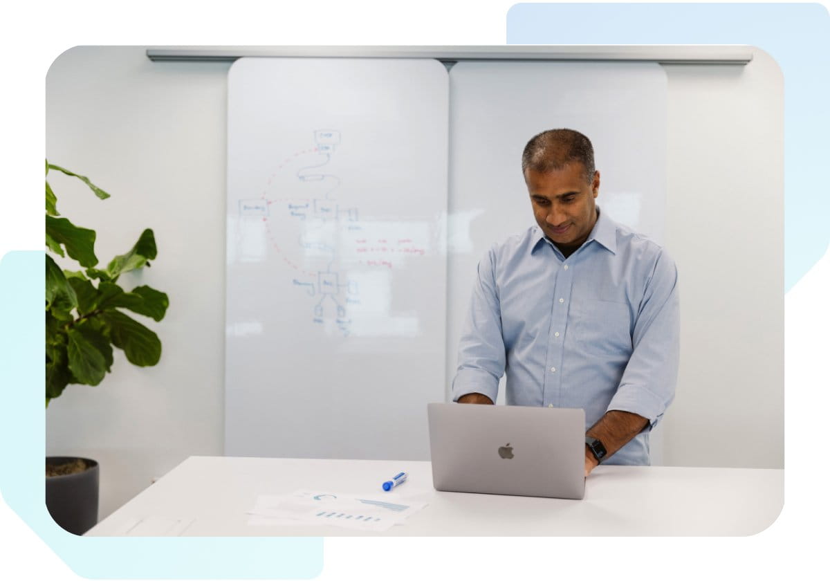 Man standing at a table looking at laptop with whiteboard behind him